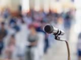 Photo focused in on a microphone, with a blurred background of people listening.