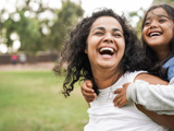 Photo of a mother and daughter outdoors, smiling, the child is being given a "piggy-back". The green park background is slightly blurred.