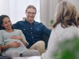 Photo of a pregnant woman, sat next to a man, both smiling, talking to a support worker (whose can be seen from behind in the image).