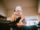 Image of a young disabled woman in a wheelchair, smiling, looking at her phone. Slightly blurred background, indoors.