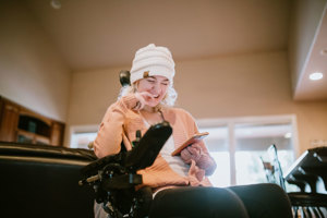Image of a young disabled woman in a wheelchair, smiling, looking at her phone. Slightly blurred background, indoors.