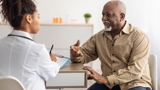 Image of an older man sat a table talking to a doctor, who is blurred and has her back to the viewer.