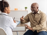Image of an older man sat a table talking to a doctor, who is blurred and has her back to the viewer.
