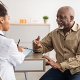 Image of an older man sat a table talking to a doctor, who is blurred and has her back to the viewer.