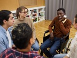 Group of people sat in a circle informally talking, some are blurred. In focus are a man in a wheelchair, a woman, and another man.