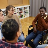 Group of people sat in a circle informally talking, some are blurred. In focus are a man in a wheelchair, a woman, and another man.