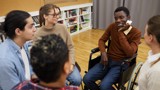 Group of people sat in a circle informally talking, some are blurred. In focus are a man in a wheelchair, a woman, and another man.