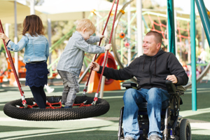 Photo of a man in a wheelchair in a park playing with his 2 children who are on a tyre swing.