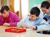 Image of 3 disabled children playing and smiling. A girl and a boy are sat at a table, playing with coloured pegboard, another boy is stood near the boy sat down, looking over his shoulder. 