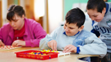 Image of 3 disabled children playing and smiling. A girl and a boy are sat at a table, playing with coloured pegboard, another boy is stood near the boy sat down, looking over his shoulder. 