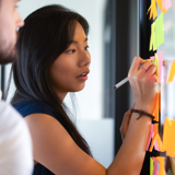 Photo focused on a woman looking at a glass board, writing notes on a post-it. There is a man standing next to her, slightly blurred.
