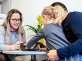 Photo of a support worker, looking at items on a desk with a child and her father.