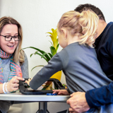 Photo of a support worker, looking at items on a desk with a child and her father.