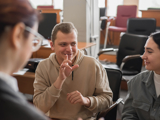 Photo of 3 people signing in a lecture hall. The man in the centre is signing, two women are communicating with him, one blurred out of focus with back to viewer.