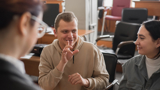 Photo of 3 people signing in a lecture hall. The man in the centre is signing, two women are communicating with him, one blurred out of focus with back to viewer.