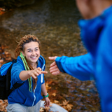 Photo focused on a young woman, smiling, outdoors walking up an incline, hand outstretched to a man reaching for her hand. He is blurred with his back to the viewer.