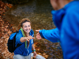 Photo focused on a young woman, smiling, outdoors walking up an incline, hand outstretched to a man reaching for her hand. He is blurred with his back to the viewer.