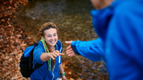Photo focused on a young woman, smiling, outdoors walking up an incline, hand outstretched to a man reaching for her hand. He is blurred with his back to the viewer.