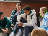 Group of 4 teenagers sitting together outside, chatting, looking at a phone