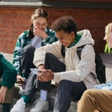 Group of 4 teenagers sitting together outside, chatting, looking at a phone