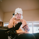 Image of a young disabled woman in a wheelchair, smiling, looking at her phone. Slightly blurred background, indoors.