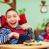 Photo of two disabled children, focused in on a boy, smiling, who is in a wheelchair, and playing with coloured bricks.