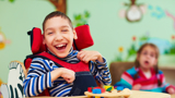 Photo of two disabled children, focused in on a boy, smiling, who is in a wheelchair, and playing with coloured bricks.