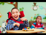 Photo of two disabled children, focused in on a boy, smiling, who is in a wheelchair, and playing with coloured bricks.