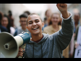 Photo focused on a smiling young woman with a megaphone in one hand and holding her other arm up. The background is blurred and of other people. 