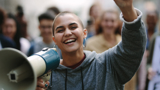 Photo focused on a smiling young woman with a megaphone in one hand and holding her other arm up. The background is blurred and of other people. 
