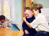 Photo focused on a man with learning disabilities, smiling, and hugging another man, also smiling. They are sat on a sofa. In the background another person is blurred.
