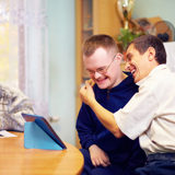 Photo focused on a man with learning disabilities, smiling, and hugging another man, also smiling. They are sat on a sofa. In the background another person is blurred.