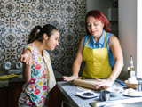 Photo in a kitchen of a young disabled woman baking with another woman, both smiling. 
