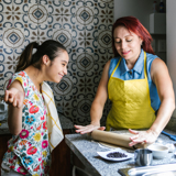 Photo in a kitchen of a young disabled woman baking with another woman, both smiling.