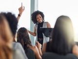 Photo of a group, with raised hands, focused in on a woman at the centre of the image happily answering questions.