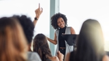 Photo of a group, with raised hands, focused in on a woman at the centre of the image happily answering questions.