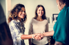 Two women meeting another woman (who has her back to the viewer), shaking hands and smiling.