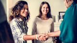 Two women meeting another woman (who has her back to the viewer), shaking hands and smiling.