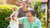 Photo focused on a middle-aged disabled woman outdoors playing with a dog, with a supporter in the background.