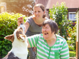 Photo focused on a middle-aged disabled woman outdoors playing with a dog, with a supporter in the background.